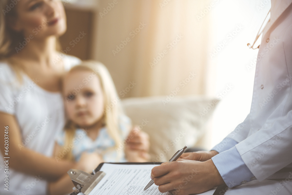 Doctor and patient. Pediatrician using clipboard while examining little girl with her mother at home. Sick and unhappy child at medical exam Doctor and patient. Pediatrician using clipboard while examining little girl with her mother at home. Sick and unhappy child at medical exam