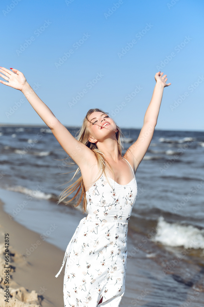 Happy blonde beautiful woman on the ocean beach standing in a white summer dress, raising hands Happy blonde beautiful woman on the ocean beach standing in a white summer dress, raising hands