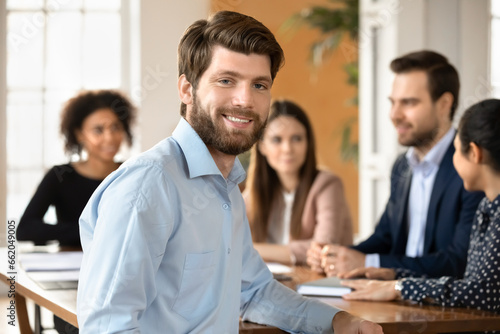 Positive handsome young project manager looking at camera, smiling, sitting at table with team of colleagues discussing cooperation, teamwork, management strategy in background