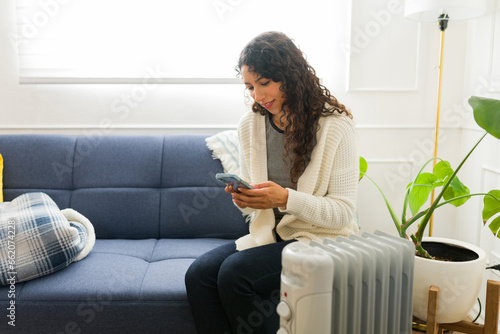 Smiling woman using the radiator heater at home relaxing with her phone