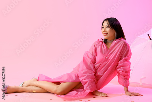 Happy fashionable young Asian woman in raincoat with umbrella sitting on pink background