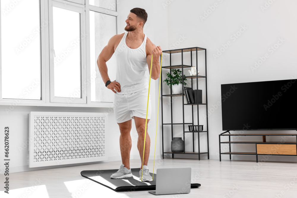 Fototapeta premium Muscular man doing exercise with elastic resistance band near laptop on mat at home