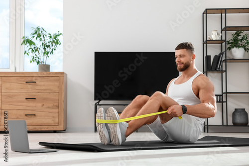 Muscular man doing exercise with elastic resistance band near laptop on mat at home © New Africa
