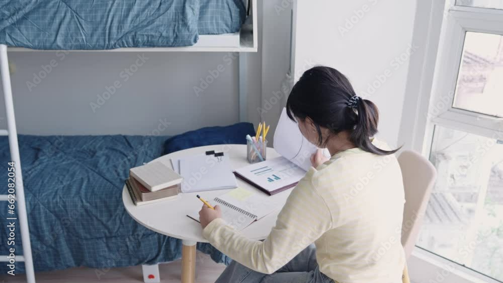 A college student does her homework writing preparing for exam test while studying paper textbook at her desk in dorm. A college student does her homework writing preparing for exam test while studying paper textbook at her desk in dorm.