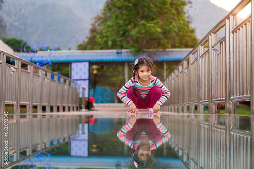 Asian child cute or kid girl happy smiling to sit squatting on skywalk bridge with reflection on clear glass floor by mountain view for vacation travel holiday relax at khao laem in vajiralongkorn dam
