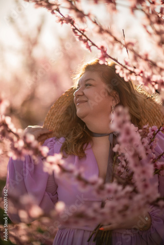 Woman blooming peach orchard. Against the backdrop of a picturesque peach orchard, a woman in a long pink dress and hat enjoys a peaceful walk in the park, surrounded by the beauty of nature.