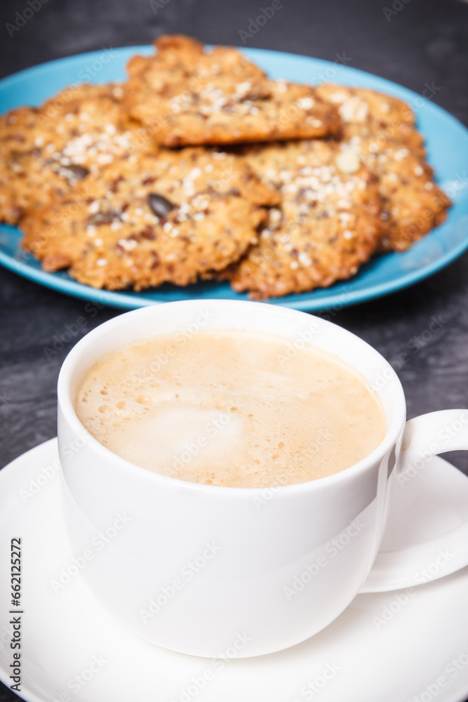 Cup of coffee with milk and fresh baked oatmeal cookies with honey and healthy seeds on blue plate. Delicious crunchy dessert Cup of coffee with milk and fresh baked oatmeal cookies with honey and healthy seeds on blue plate. Delicious crunchy dessert
