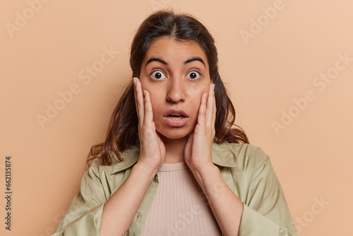 Portrait of scared Iranian woman keeps hands on cheeks stares with bugged eyes and feels speechless wears casual shirt cannot believe in shocking news isolated on brown background. Omg what relevation photo
