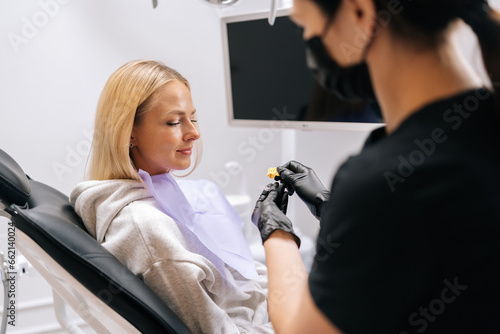 Side view of unrecognizable female orthodontist doctor showing aligners or kappa for teeth alignment to young woman patient, sitting on dental chair in dentistry clinic. Concept of orthodontics.