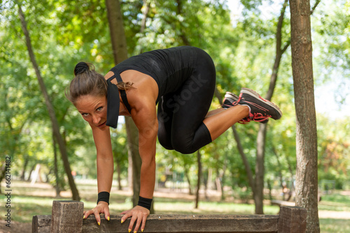 An athletic woman showcases her agility, leaping over an outdoor wooden barrier in a park.