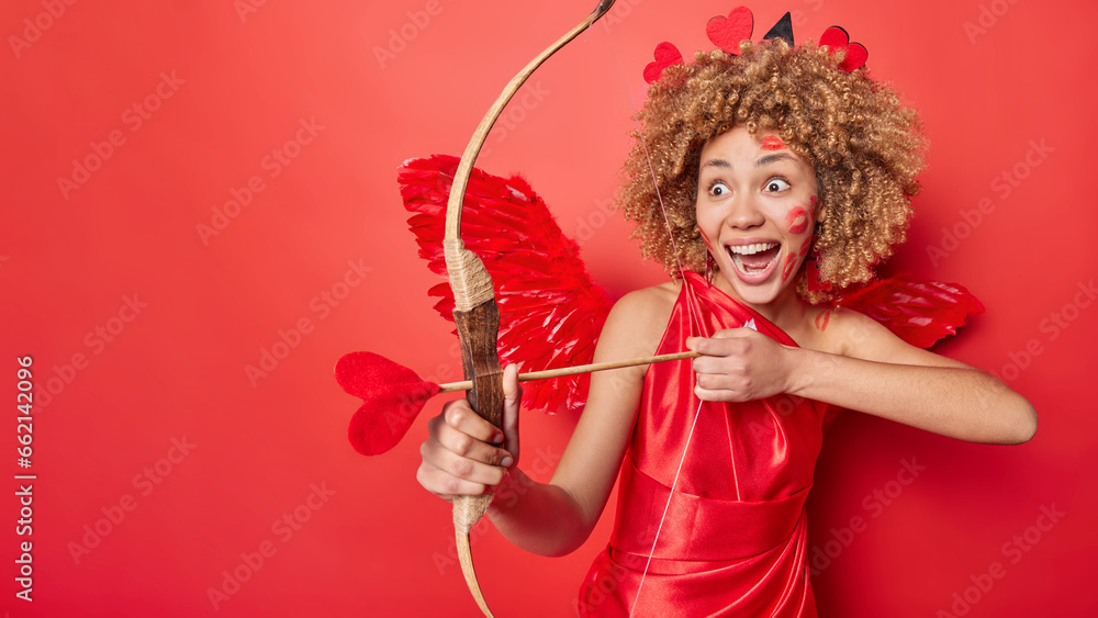Surprised curly female cupid targets at someone with bow tries to match people dressed in long dress poses against vivid red background with copy space for your text. Love and romance concept Surprised curly female cupid targets at someone with bow tries to match people dressed in long dress poses against vivid red background with copy space for your text. Love and romance concept