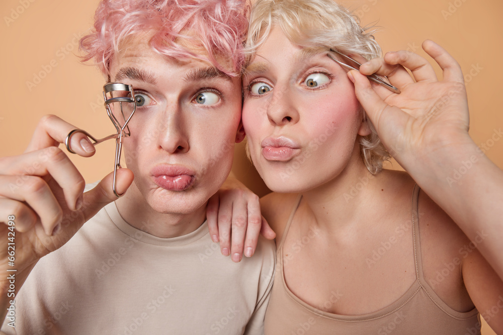Beauty procedures and wellness concept. Funny woman and man use eyelashes curler and plucker want to look beautiful keep lips folded dressed in casual t shirts isolated over brown background.