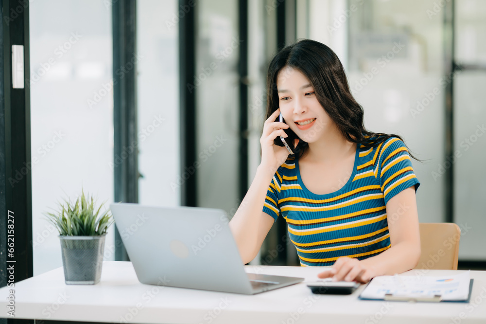 Smiling Asian woman talking on the phone with a customer Young positive female accountant using smartphone talking to team at her desk relaxing with mobile Smiling Asian woman talking on the phone with a customer Young positive female accountant using smartphone talking to team at her desk relaxing with mobile