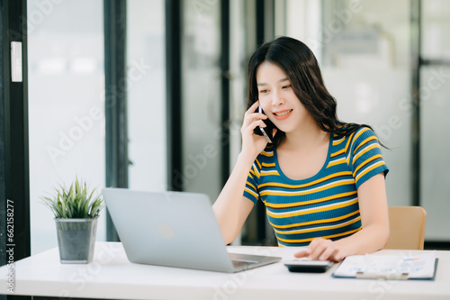 Smiling Asian woman talking on the phone with a customer Young positive female accountant using smartphone talking to team at her desk relaxing with mobile