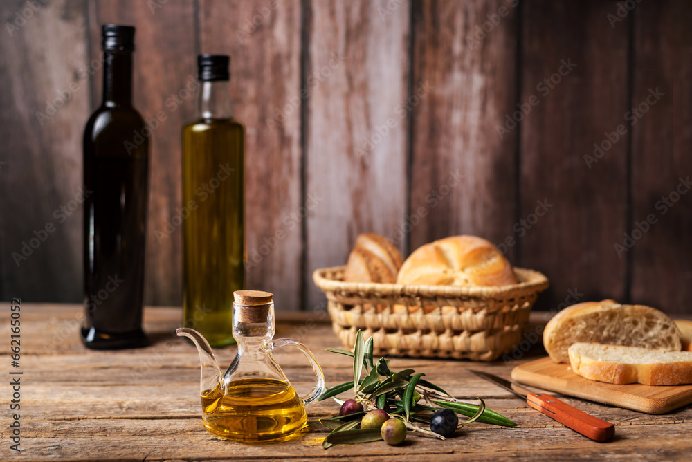 Oil jug with virgin olive oil next to an olive branch with olives, in the background bread and some bottles of olive oil, rustic background.