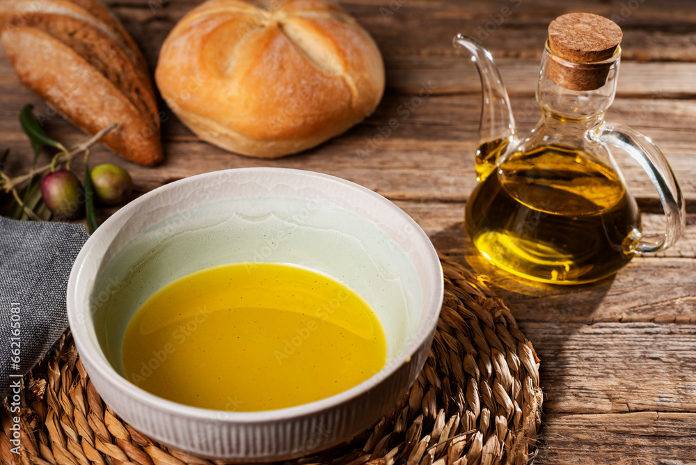 Bowl with olive oil next to an oil can and some bread rolls, with a rustic background. Bowl with olive oil next to an oil can and some bread rolls, with a rustic background.
