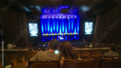 Young couple sits in chairs in half-empty concert hall before show starts, stage illuminated with blue lights. They using smartphone, taking selfies or checking social networks