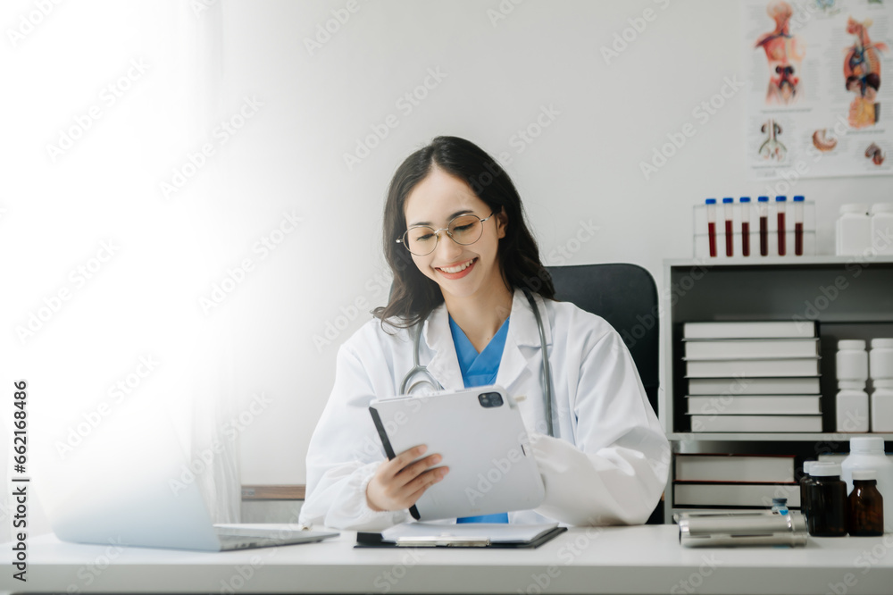 Confident young Asian female doctor in white medical uniform sit at desk working on computer. Smiling use laptop write in medical journal Confident young Asian female doctor in white medical uniform sit at desk working on computer. Smiling use laptop write in medical journal