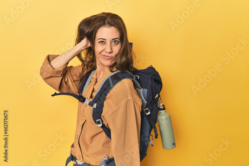 Middle aged woman prepped for hiking, yellow studio shot touching back of head, thinking and making a choice.