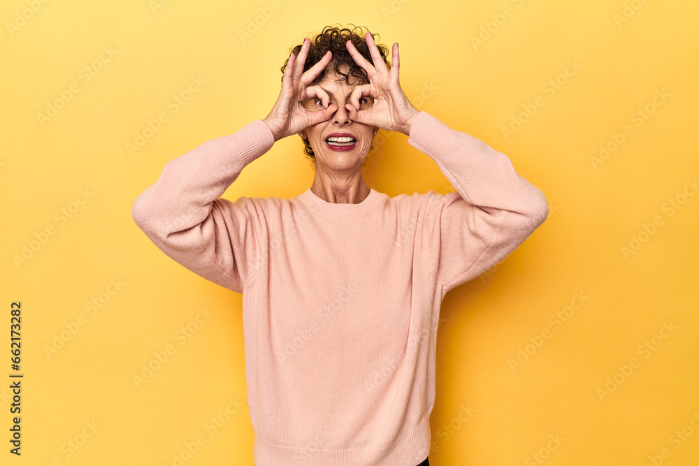 Mid-aged caucasian woman on vibrant yellow showing okay sign over eyes Mid-aged caucasian woman on vibrant yellow showing okay sign over eyes