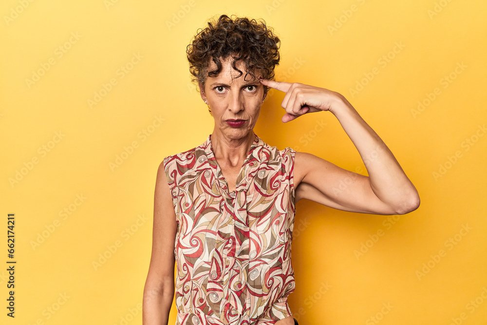 Mid-aged caucasian woman on vibrant yellow pointing temple with finger, thinking, focused on a task. Mid-aged caucasian woman on vibrant yellow pointing temple with finger, thinking, focused on a task.