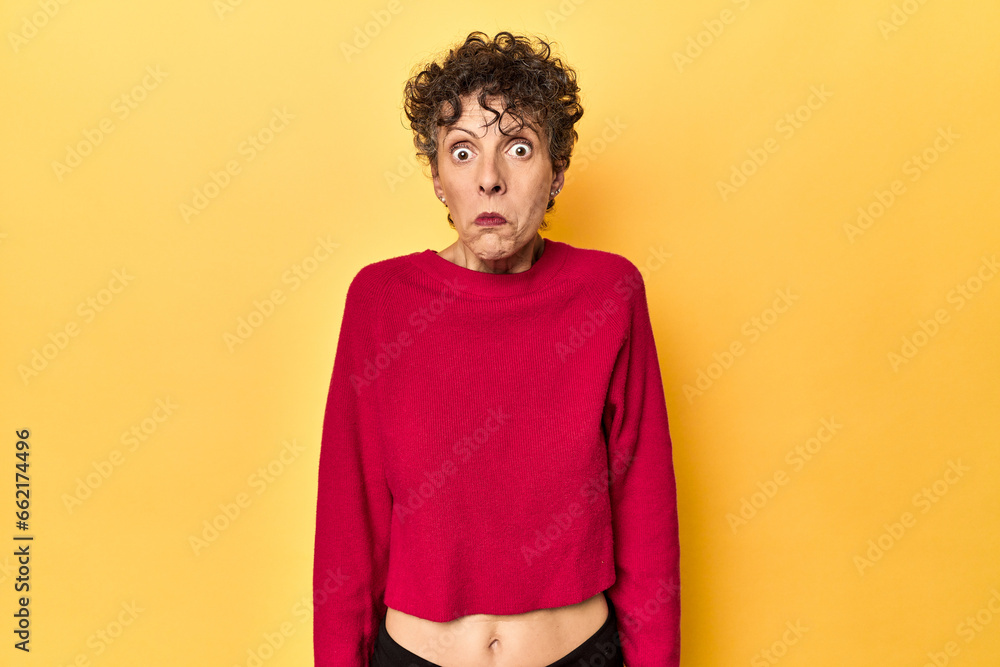 Mid-aged caucasian woman on vibrant yellow shrugs shoulders and open eyes confused. Mid-aged caucasian woman on vibrant yellow shrugs shoulders and open eyes confused.