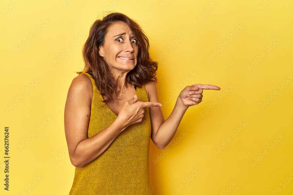Middle-aged woman on a yellow backdrop shocked pointing with index fingers to a copy space. Middle-aged woman on a yellow backdrop shocked pointing with index fingers to a copy space.