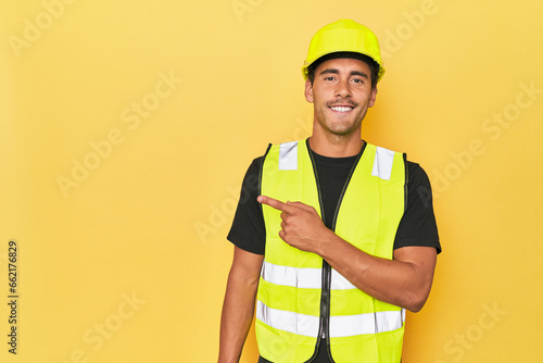 Latino worker in yellow vest and helmet smiling and pointing aside, showing something at blank space.