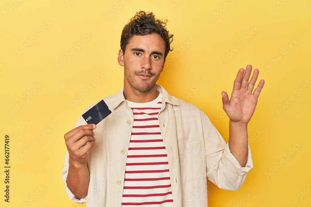 Man holding credit card on yellow smiling cheerful showing number five with fingers. Man holding credit card on yellow smiling cheerful showing number five with fingers.