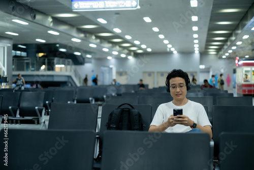 Young Asian millenael man sitting in air port or rail station holding smartphone ready for trip travel with black backpack and headphone.