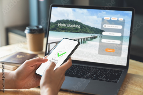Hand using laptop and booking hotel on website for vacation after a long work and payment completed message on mobile, Summer holiday concept.