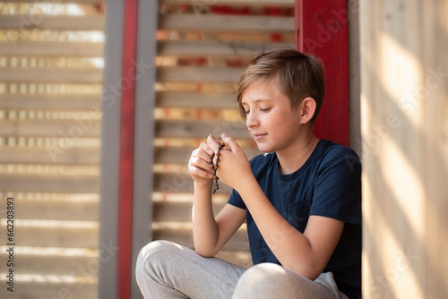 An 11 year old Catholic boy reads the rosary prayer, holds a wooden rosary with 10 beads in his hands. portrait of a boy with a wooden Catholic rosary during prayer.	
 photo