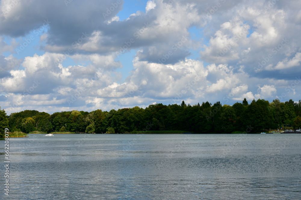 View of a small urban beach surrounded from all sides with lush forests, moors, and shrubbery, with a sandy coast, wooden platforms and piers, as well as boats swimming on the lake or river in summer