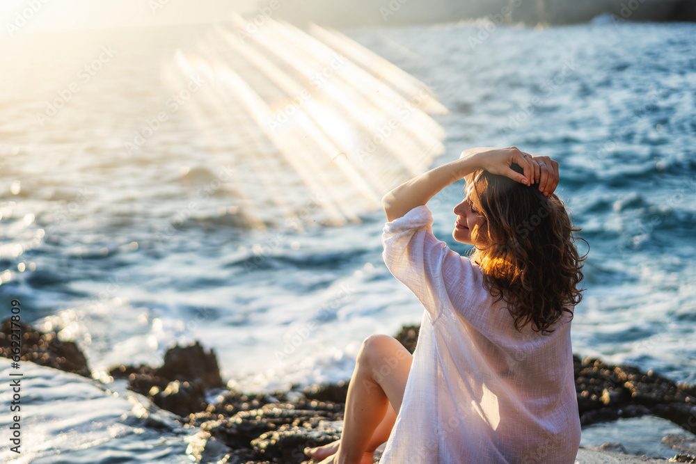 Young cheerful relaxed woman in a white shirt enjoying a wonderful moment of life while sitting on the seashore at sunset