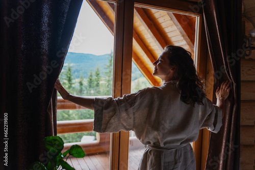 Young caucasian woman opening curtains in a wooden chalet cabin in the mountains. Comfortable rest in a log house in nature conept