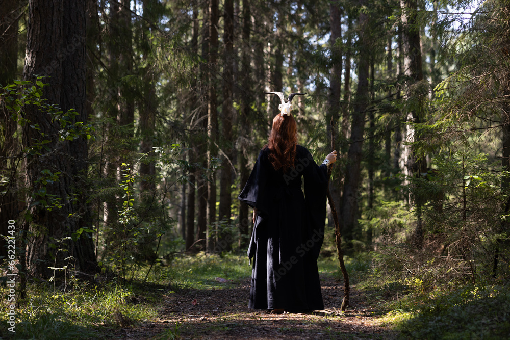Fototapeta premium A forest girl walks along the road with her back to us. She has a staff in her hand