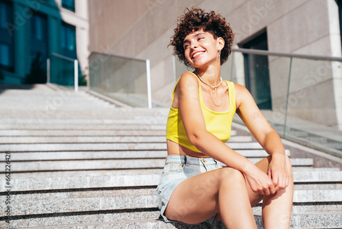 Young beautiful smiling hipster woman in trendy summer clothes. Carefree woman with curls hairstyle, posing on the street at sunny day. Positive model outdoors. Cheerful and happy, sits at stairs