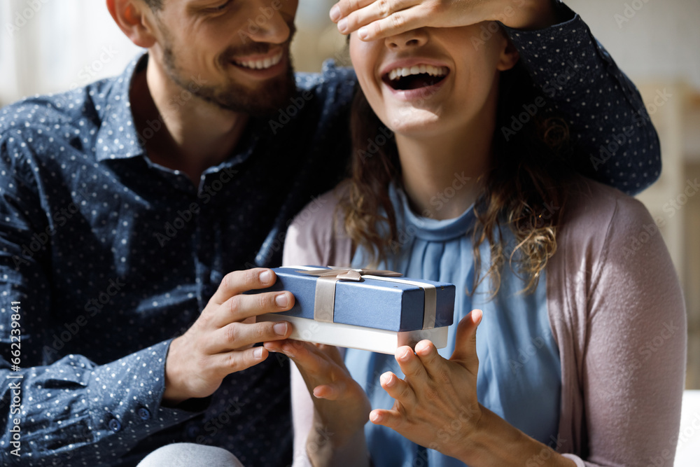 Fototapeta premium Excited dating couple celebrating Valentines day, 8 march, anniversary, birthday. Happy husband giving surprise gift box to cheerful wife with closed shut eyes. Love, relationship concept. Close up