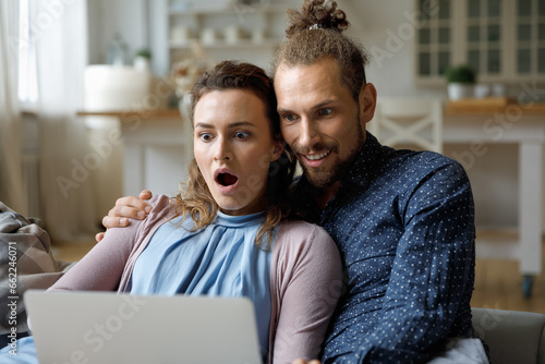Shocked happy young married couple getting email message with surprising good news, looking at laptop computer with excitement, winning money prize, learning about luck, online sale, discount