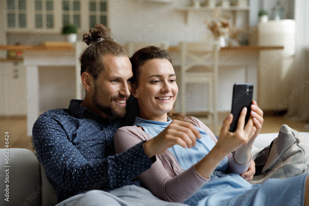 Happy sweet young couple taking selfie picture on mobile phone, watching live media content, talking on video call, using online app on digital gadget, relaxing on couch, enjoying leisure at home