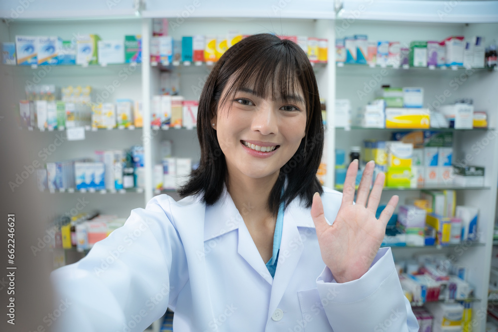 Young asian female pharmacist holding phone to selfie with smiling at pharmacy counter.