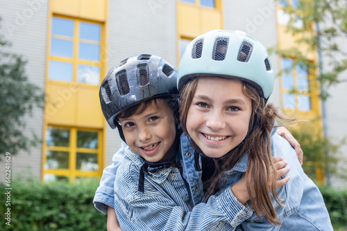 Close-up portrait of two children, boy and girl, hugging, looking at the camera, smiling in helmets, ready to go roller skating or cycling in their free time from lessons in the school yard on break. © SistersStock