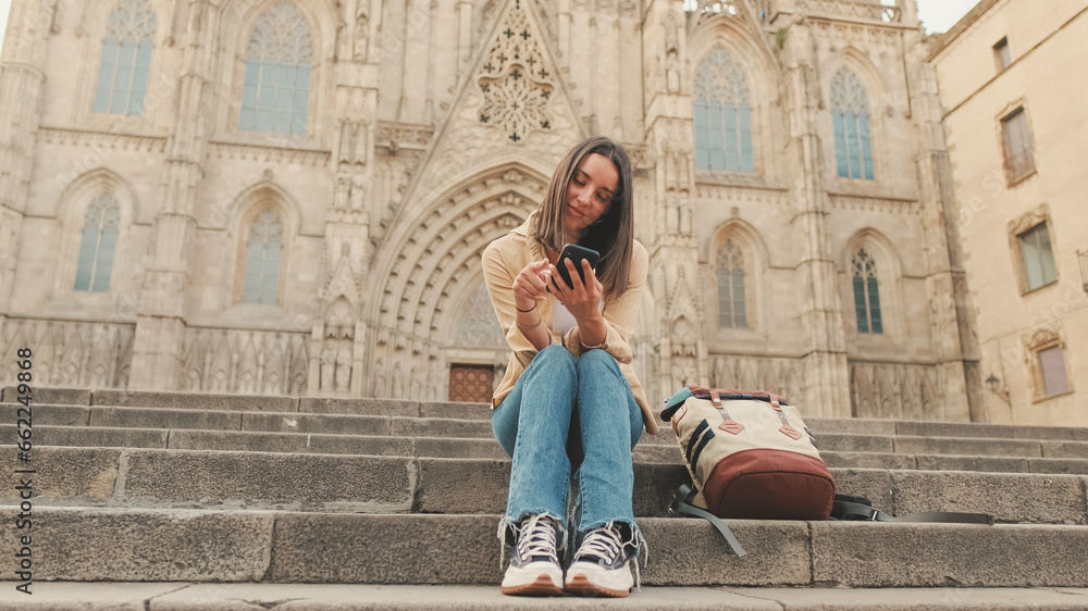Happy traveler girl using mobile phone while sitting on the steps of an old building in the historic part of an old European city - obrazy, fototapety, plakaty 
