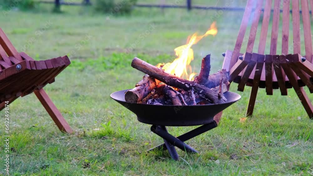 Metal fire bowl with burning wood in the yard and garden rack lounge chair. Safe decorative fire pit, Picnic area in the garden Metal fire bowl with burning wood in the yard and garden rack lounge chair. Safe decorative fire pit, Picnic area in the garden