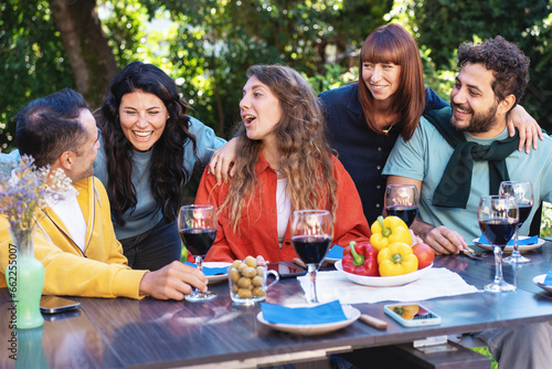 Joyful Friends Sharing a Meal Outdoors: A lively group of five friends laugh and bond over a table filled with wine and fresh produce in a garden setting.