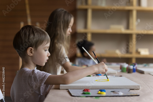 Beautiful kids painting pictures on canvas sit at table in art-studio. Group classes of art for little children to develop talent, explore their imagination and express themselves in creative ways