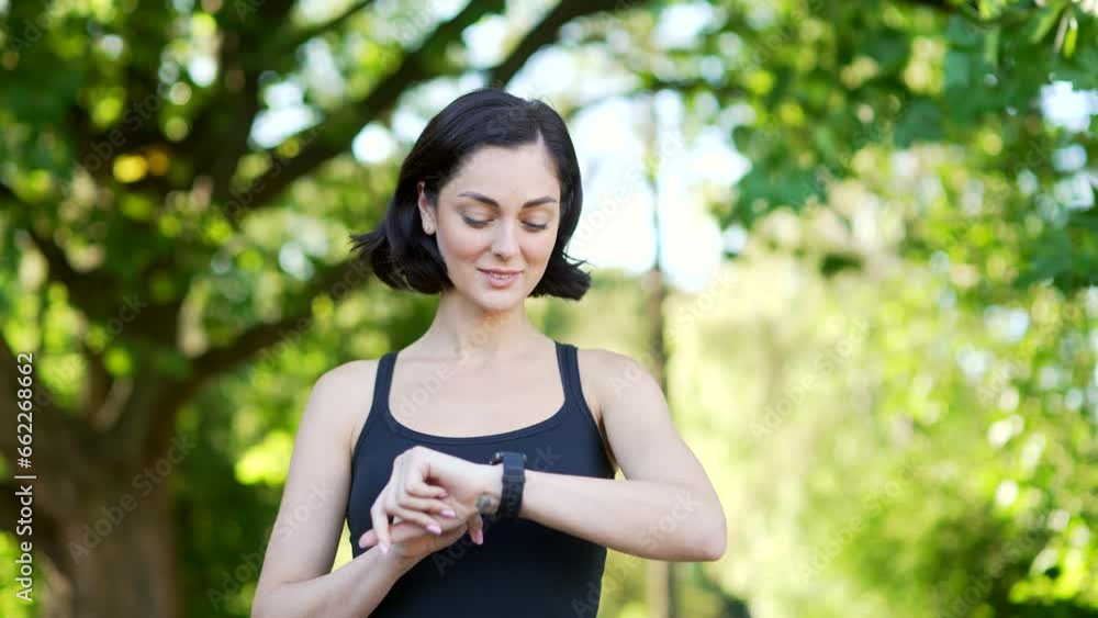 A young cute female runner stands in an urban city park and looking smart watch. Sportswoman using fitness tracker. Fit female checking result is satisfied with indicators of training, rejoices succes A young cute female runner stands in an urban city park and looking smart watch. Sportswoman using fitness tracker. Fit female checking result is satisfied with indicators of training, rejoices succes