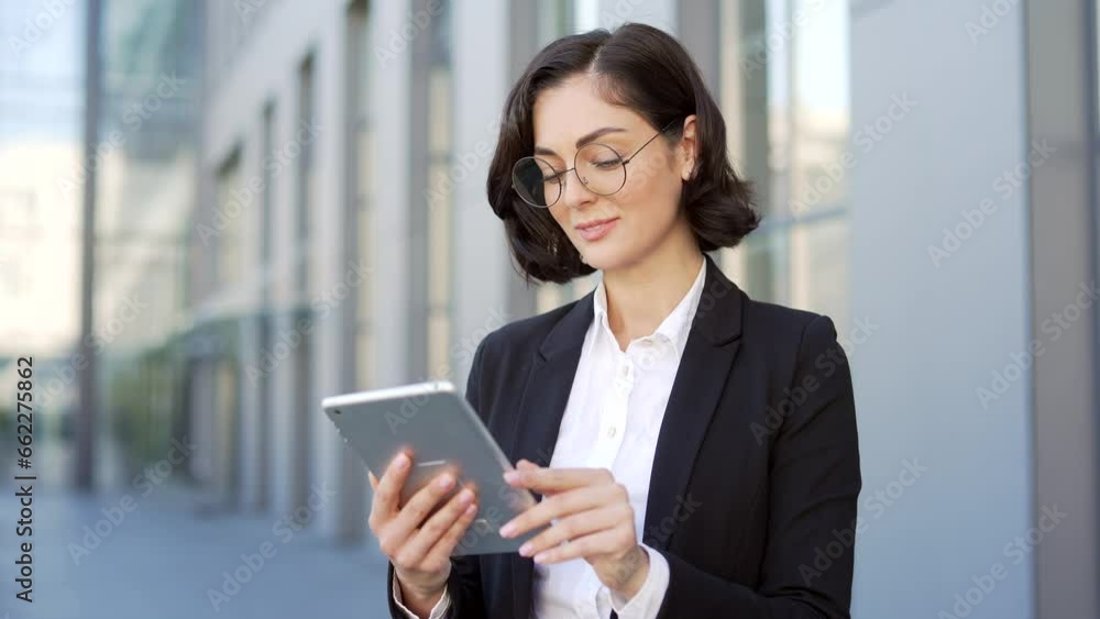 Portrait of a young smiling businesswoman standing on the street near an office building using a tablet. A brunette woman in a formal suit and glasses looking at the camera. Headshot of female manager Portrait of a young smiling businesswoman standing on the street near an office building using a tablet. A brunette woman in a formal suit and glasses looking at the camera. Headshot of female manager
