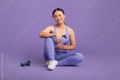 happy asian woman holding bottle with water, sitting on floor over purple studio background