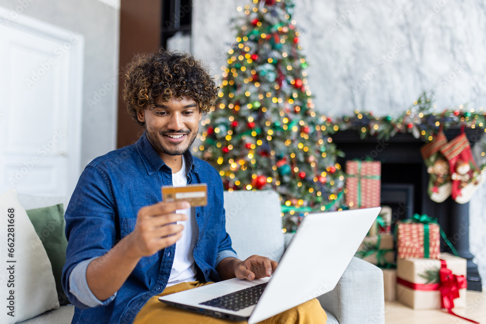 Man is sitting on a sofa in the living room of the house for Christmas, hispanic smiling, satisfied, choosing gifts and goods online in an online store, holding a bank credit card near tree. Man is sitting on a sofa in the living room of the house for Christmas, hispanic smiling, satisfied, choosing gifts and goods online in an online store, holding a bank credit card near tree.
